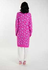 Cárdigan de punto largo y rosa con textura suave y patrón de manchas moradas claras, llevado sobre pantalones blancos y tacones rosas, vista desde la espalda.