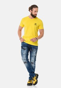 Gelbes kurzärmliges V-Ausschnitt-T-Shirt mit schwarzem Logo, kombiniert mit distressed blauen Jeans mit mehreren Taschen und gemusterten Akzenten sowie schwarz-gelben Sneakern.