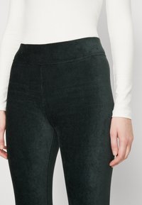 Donna che indossa leggings neri in corda a vita alta e una maglietta bianca a maniche lunghe, con le mani appoggiate ai fianchi su uno sfondo grigio.