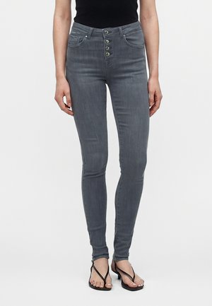 Kvinde iført grå skinny jeans med høj talje og fire knapper foran samt sorte hælede sandaler med stropper, stående mod en hvid baggrund.