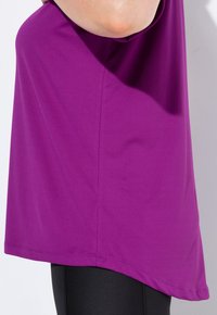 Kvinde iført en løs lilla top og sorte teksturerede leggings, vist fra siden ved midt-torso niveau mod en ensfarvet baggrund.