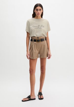 Vrouw die een beige T-shirt met tekst draagt, een high-waist tan korte broek met een zwarte riem en zwarte sandalen, staand tegen een effen witte achtergrond.