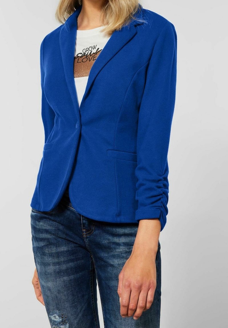 Femme portant un blazer ajusté bleu roi, un t-shirt blanc avec un motif graphique et un jean bleu foncé usé, les mains relâchées sur les côtés.