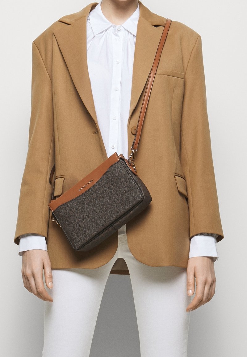 Personne portant un blazer camel et une chemise blanche tenant un sac bandoulière marron et gris foncé avec une sangle en cuir.