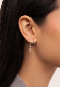 Pendientes de aro de plata con acentos coloridos y un diseño colgante que presenta tres piedras transparentes, exhibidos en una oreja.