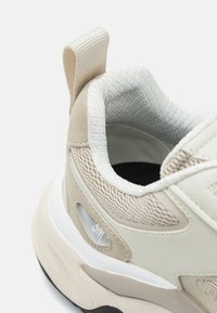 Beige sportcipő sima bőr és texturált mesh kombinációjával. Lekerekített forma, húzófül a saroknál, és kontrasztos fekete talp.