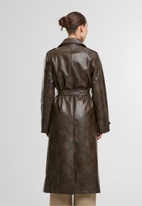 Brązowy płaszcz trench z sztucznej skóry z paskiem w talii, długimi rękawami i gładką fakturą, bez widocznych elementów metalowych ani zdobień.