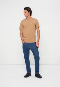 T-shirt beige à manches courtes avec un petit logo jaune, associé à un jean en denim bleu et des chaussures noires, sur fond blanc uni.