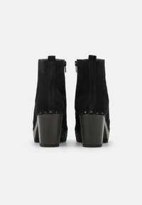 Un par de botines de ante negros con tacones de bloque y cremalleras traseras, mostrados desde atrás sobre un fondo blanco.