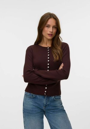 Kvinde med langt brunt hår iført en mørkebrun knappet cardigan og blå jeans, stående med armene krydset mod en ensfarvet baggrund.