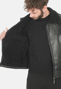 Giubbotto di pelle nera con foderatura in lana texturizzata, chiusura con zip e dettaglio con fibbia al colletto, indossato sopra un maglione nero a treccia.