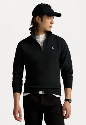 Sweatshirt negru cu guler cu fermoar, dintr-un amestec de bumbac, cu broderie albă a logo-ului, purtat peste un tricou alb, asortat cu pantaloni negri și o șapcă neagră.