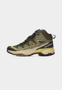 Πέδιλο πεζοπορίας μεσαίου ύψους σε ελιά πράσινο και μπεζ, με κίτρινες λεπτομέρειες, μαύρη σόλα και λογότυπο Salomon στην πλευρά της φτέρνας.