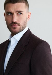 Burgunderfarbener Wollmischblazer mit Reverskragen, einer einzelnen Brusttasche und einem weißen Hemd darunter, das eine glatte Textur zeigt.