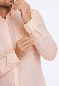 Camisa de botones en color durazno claro con un bolsillo, tela texturizada y puños ajustables. Presenta un cuello clásico y un patrón sutil.