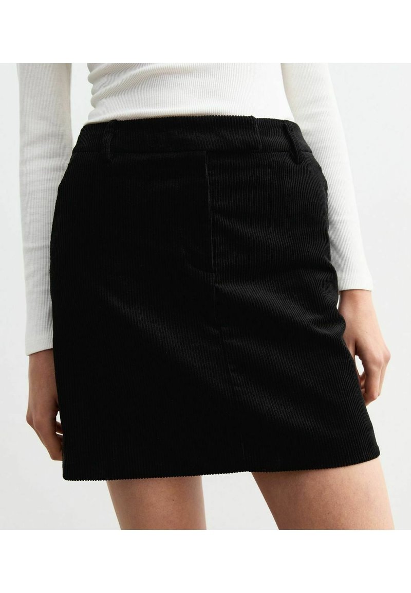 Femme portant une mini-jupe en velours côtelé noire et un haut blanc à manches longues côtelé, debout les bras relâchés le long du corps.