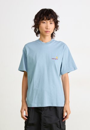 Persona con capelli ricci che indossa una T-shirt Carhartt azzurra chiara e pantaloni cargo neri, in piedi contro uno sfondo semplice.