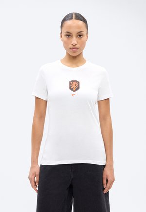Frau steht frontal, trägt ein weißes T-Shirt mit orangefarbenem Löwenwappen und Nike-Logo, kombiniert mit schwarzer Hose, vor einfarbigem Hintergrund.