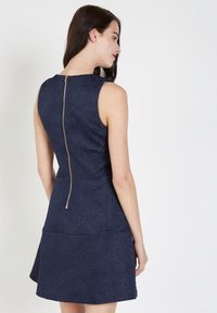 Vestido azul marino con patrón floral texturizado, parte superior ajustada, falda acampanada y cremallera trasera visible. Tejido suave y diseño sin mangas.