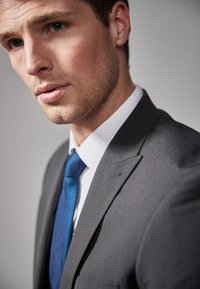 Veste de costume grise avec revers en pointe, chemise blanche et cravate en tissu texturé bleu. Présente des détails de couture subtils sur le revers et une texture de tissu lisse.