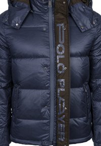 Námořnická puffer bunda s lesklou texturou, vybavená předním zipem, patentkami a velkým logem na boku v kontrastní hnědé barvě.