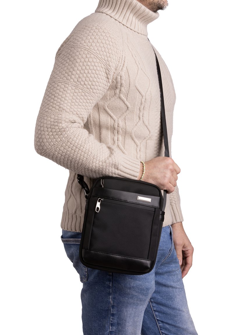 Borsa a tracolla nera con superficie testurizzata, tasca frontale con zip e tracolla regolabile, indossata da una persona con un maglione lavorato a maglia beige.