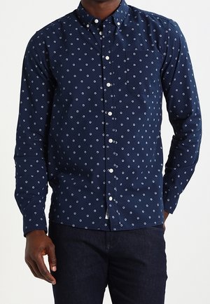 Homme portant une chemise bleu marine à boutons avec un motif circulaire blanc et un pantalon bleu foncé, debout avec une main dans la poche.