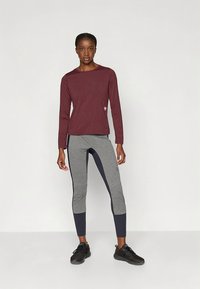 Bordeauxrode longsleeve gecombineerd met grijze en marineblauwe legging, met een aansluitend ontwerp en contrasterende panelen, gedragen met zwarte sportschoenen.