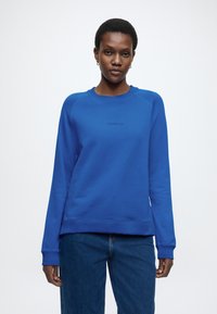 Kék, kerek nyakú pulóver hosszú ujjakkal, bordázott mandzsettákkal és szegéllyel, mellkasán kis logóval. Puha, sima tapintású anyag.