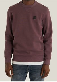 Felpa color bordeaux con scollo rotondo e polsini a costine. Presenta una piccola toppa con logo nero sul petto. Il materiale sembra morbido e strutturato.