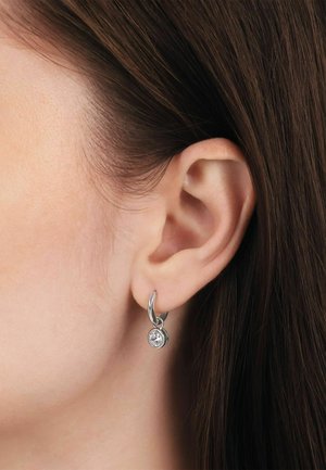 Primer plano de la oreja de una mujer con cabello castaño que lleva un pequeño aro de plata con un único colgante de piedra preciosa transparente.