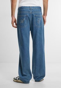 Pantalones de mezclilla azul con un ajuste relajado y piernas anchas, que presentan dos bolsillos traseros adornados con letras bordadas "A" y "C."