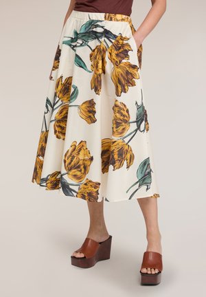 Mid-lang hvid nederdel med stort gult blomsterprint, kombineret med brune kile-sandaler og en brun top stoppet ind. Hænder i nederdelens lommer.