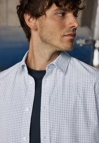 Wit-blauw geruit overhemd met een klassieke kraag, knopen aan de voorkant en een gladde textuur, gedragen over een donker T-shirt.