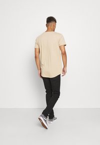 T-shirt beige à manches courtes, avec un ourlet arrondi. Porté avec un jean noir skinny et des baskets grises avec des accents blancs, vu de dos.