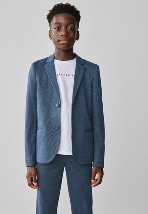 Veste de costume texturée bleue avec deux boutons, revers crantés et poches latérales, portée sur un t-shirt blanc avec un texte noir.