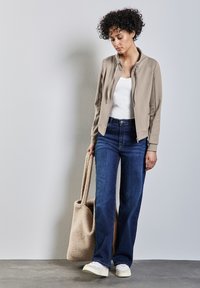 Giacca bomber beige con zip, canotta bianca, pantaloni a gamba larga blu scuro e sneakers off-white; tiene una borsa a tracolla beige e soffice.