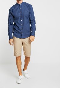 Mörkblå långärmad knäppskjorta med mandarin-krage, kombinerad med khaki-shorts och vita sneakers, vilket visar en avslappnad outfit.