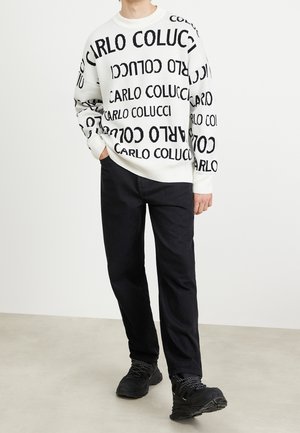 Un pull blanc avec un motif de texte "CARLO COLUCCI" en noir, associé à un pantalon noir et des chaussures noires robustes, présentant un tissu texturé et une coupe décontractée.