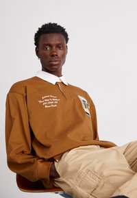 Brun långärmad skjorta med vit krage, med broderad text och en grafisk logotyp. Modellen sitter och har på sig khakifärgade byxor.
