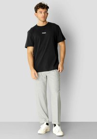 Svart bomull T-skjorte med en liten hvit tekstlogo, paret med lysegrå, strukturerte bukser og hvite joggesko med svarte detaljer.