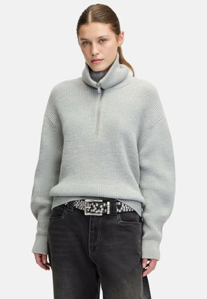 Hellgrauer, gestrickter Pullover mit hohem, umgeschlagenem Kragen und einem kurzen Reißverschluss. Hat lockere Ärmel und einen gerippten Saum, kombiniert mit schwarzen Jeans und einem Nieten-Gürtel.