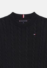 Pull en tricot noir avec un col rond, des poignets et un ourlet côtelés. Petite plaque logo rouge, blanc et bleu sur la poitrine.
