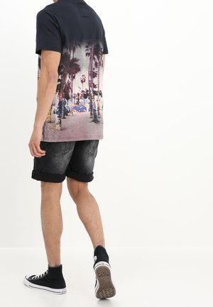 Mężczyzna idący, ubrany w czarne trampki za kostkę, podwinięte ciemne szorty z denimu oraz czarny T-shirt z nadrukiem plaży i palmy.