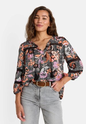 Blouse noire à motifs floraux vifs orange et rose, décolleté en V, manches bouffantes longues et accents noirs. Associée à un jean gris clair et une ceinture marron.