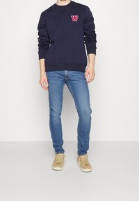 Man med marinblå sweatshirt med röd emblem, ljusblå slim jeans och beige sneakers, stående mot en enkel vit bakgrund.