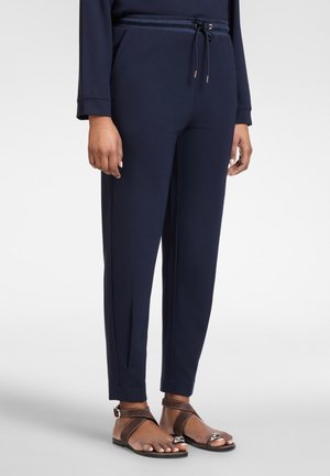 Marineblaue, schmal zulaufende Hose mit Kordelzugbund, getragen mit braunen Leder-Sandalen mit offenem Zehenbereich bei einer stehenden Person.