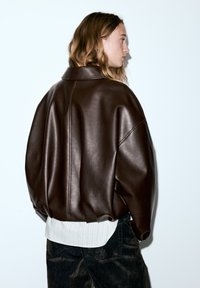 Veste oversize en cuir marron avec un large col, des épaules arrondies et des coutures prononcées, portée sur une chemise blanche à rayures et un jean foncé.