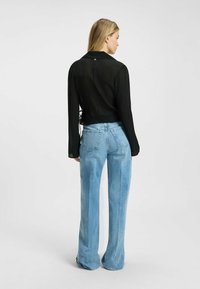 Sort gennemsigtig bluse med lange ærmer, krave og bånddetalje bagpå, parret med lys blå højtaljede flared jeans, vist fra bagvenden.