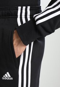 Czarne dresowe spodnie Adidas mają trzy białe paski po bokach, elastyczny pas, kieszeń boczną i teksturowany materiał.